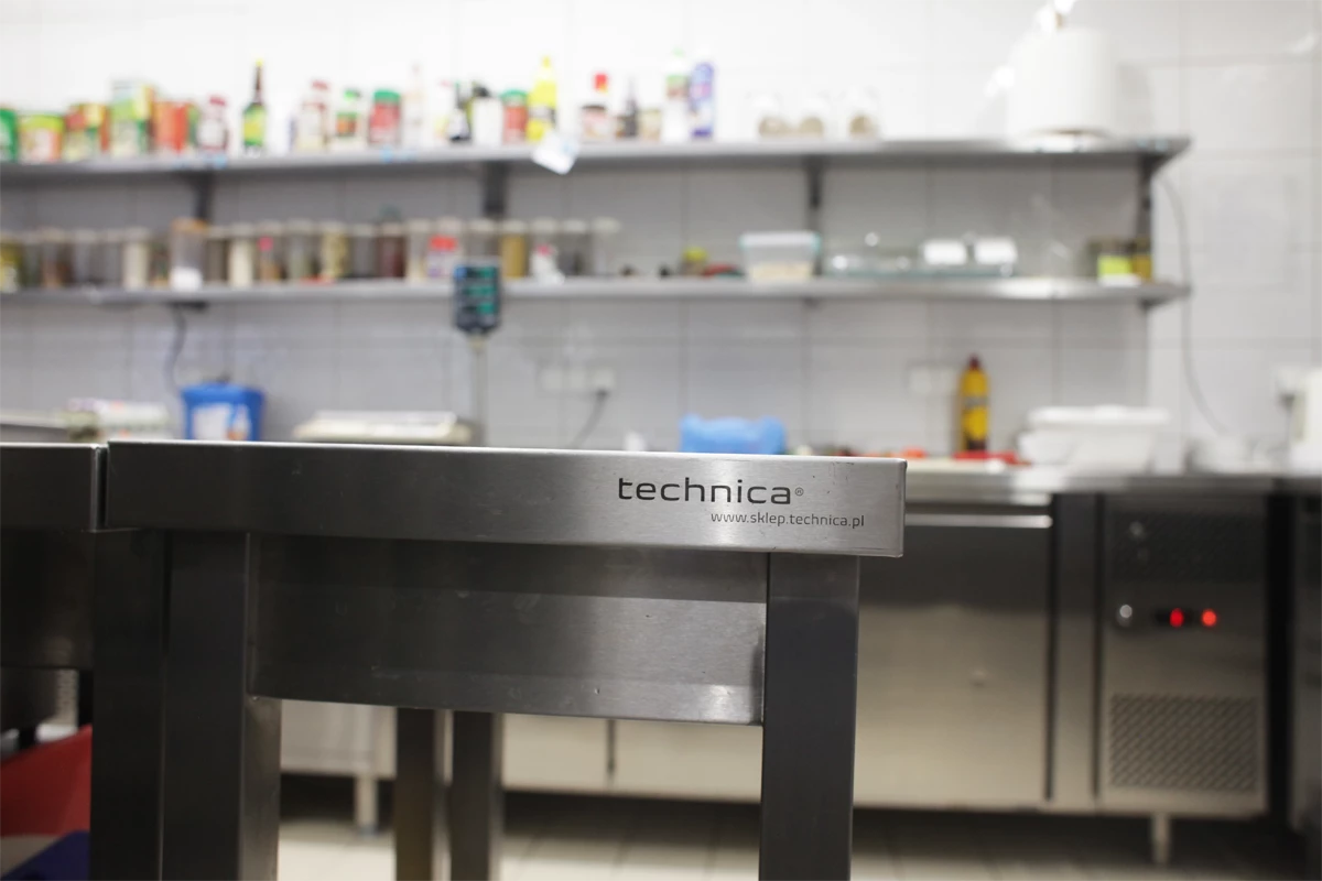 Fragment stołu roboczego ze stali nierdzewnej z detalem na logo producenta – technica, w profesjonalnej kuchni gastronomicznej.