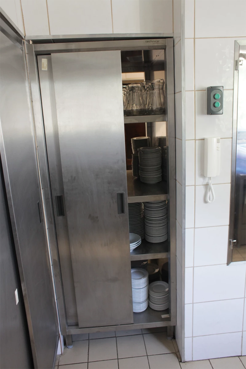 Szafa przelotowa ze stali nierdzewnej z przesuwnymi drzwiami, wypełniona talerzami i szklanymi dzbankami, zamontowana w kuchni gastronomicznej.