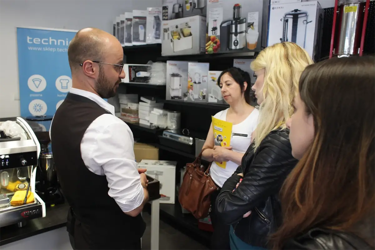 Kuchenka gazowa z sześcioma palnikami i stanowiskiem roboczym technica w profesjonalnej kuchni gastronomicznej. W tle regały i półki z naczyniami oraz akcesoriami kuchennymi.