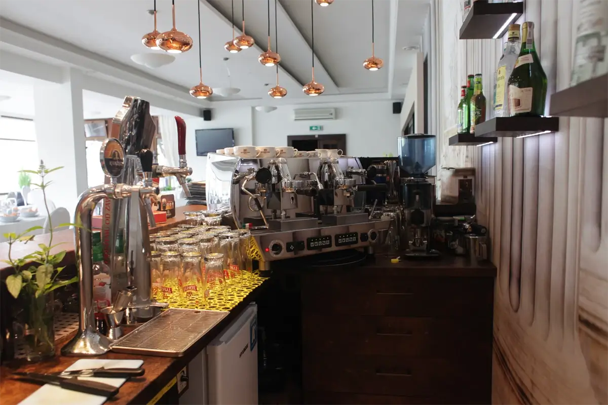 Stanowisko barowe z ekspresami do kawy, szklankami, nalewakami do piwa i butelkami alkoholu na podświetlanych półkach, w nowoczesnym, jasnym wnętrzu restauracji.