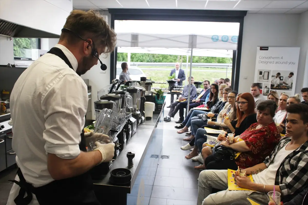 Wnętrze nowoczesnej kuchni gastronomicznej z centralnym blokiem roboczym, kuchenkami gazowymi, dużym garnkiem, piecem konwekcyjnym i regałami ze stali nierdzewnej pod wyciągiem wentylacyjnym.