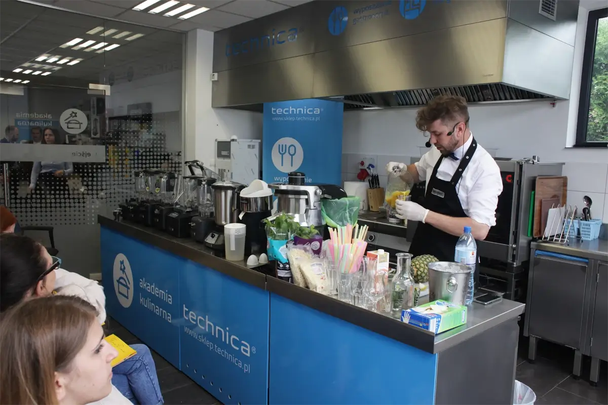 Stanowisko robocze w kuchni gastronomicznej z blatami i półkami ze stali nierdzewnej, zmywarką, lodówką, naczyniami i akcesoriami kuchennymi, na tle białych płytek ściennych.