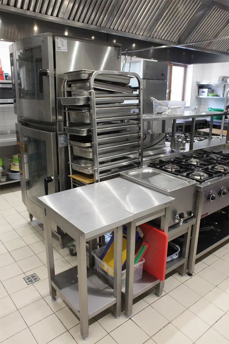 Kuchnia gastronomiczna z piecami konwekcyjnymi, wózkiem transportowym ze stali nierdzewnej z pojemnikami GN, blatami roboczymi ze stali nierdzewnej oraz zestawem desek HACCP do krojenia.