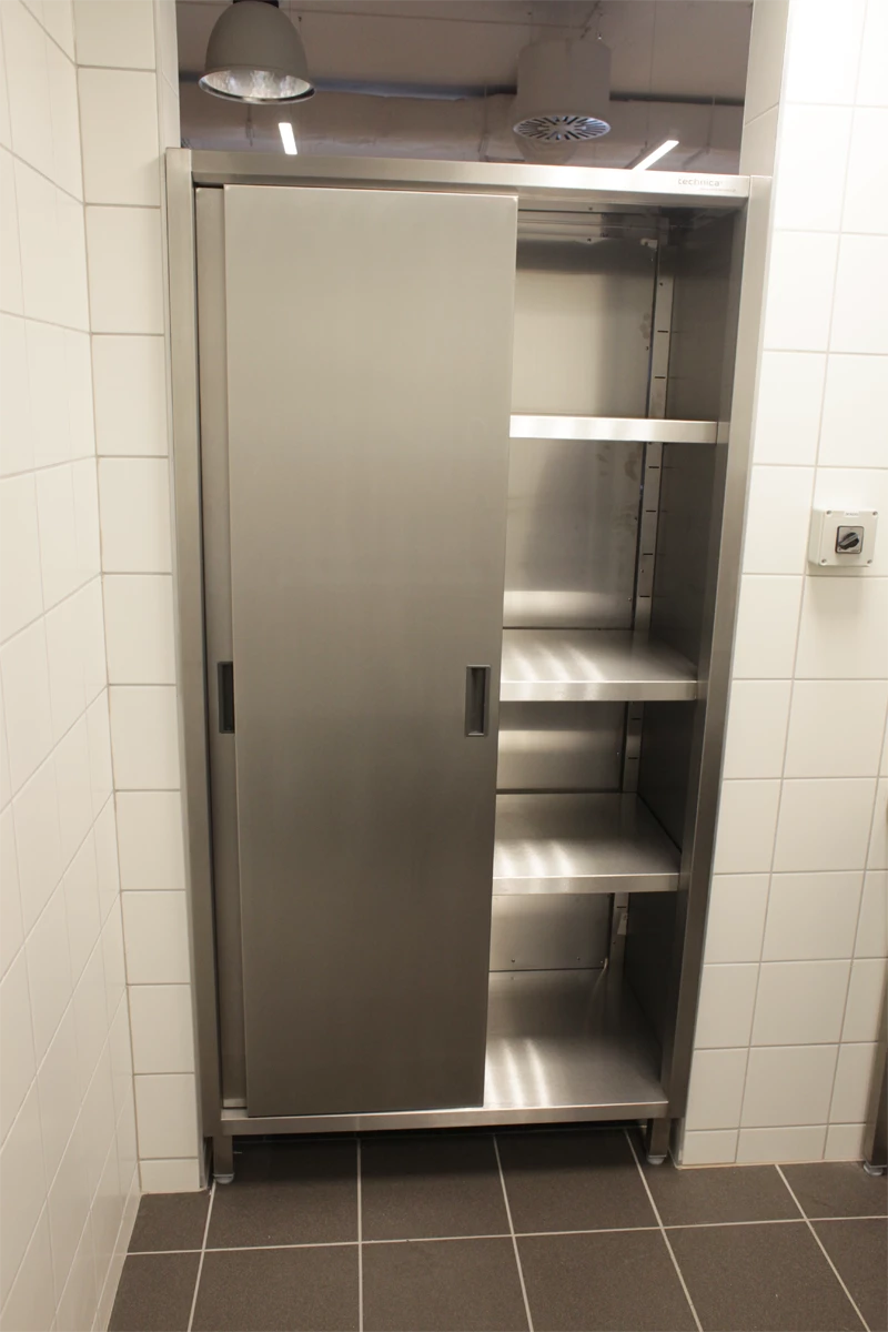 Szafa przelotowa ze stali nierdzewnej, z drzwiami przesuwnymi, umieszczona w przestrzeni z białymi kafelkami na ścianach. Szafka ma półki, a wnętrze jest puste. W tle widać elementy oświetlenia sufitowego.