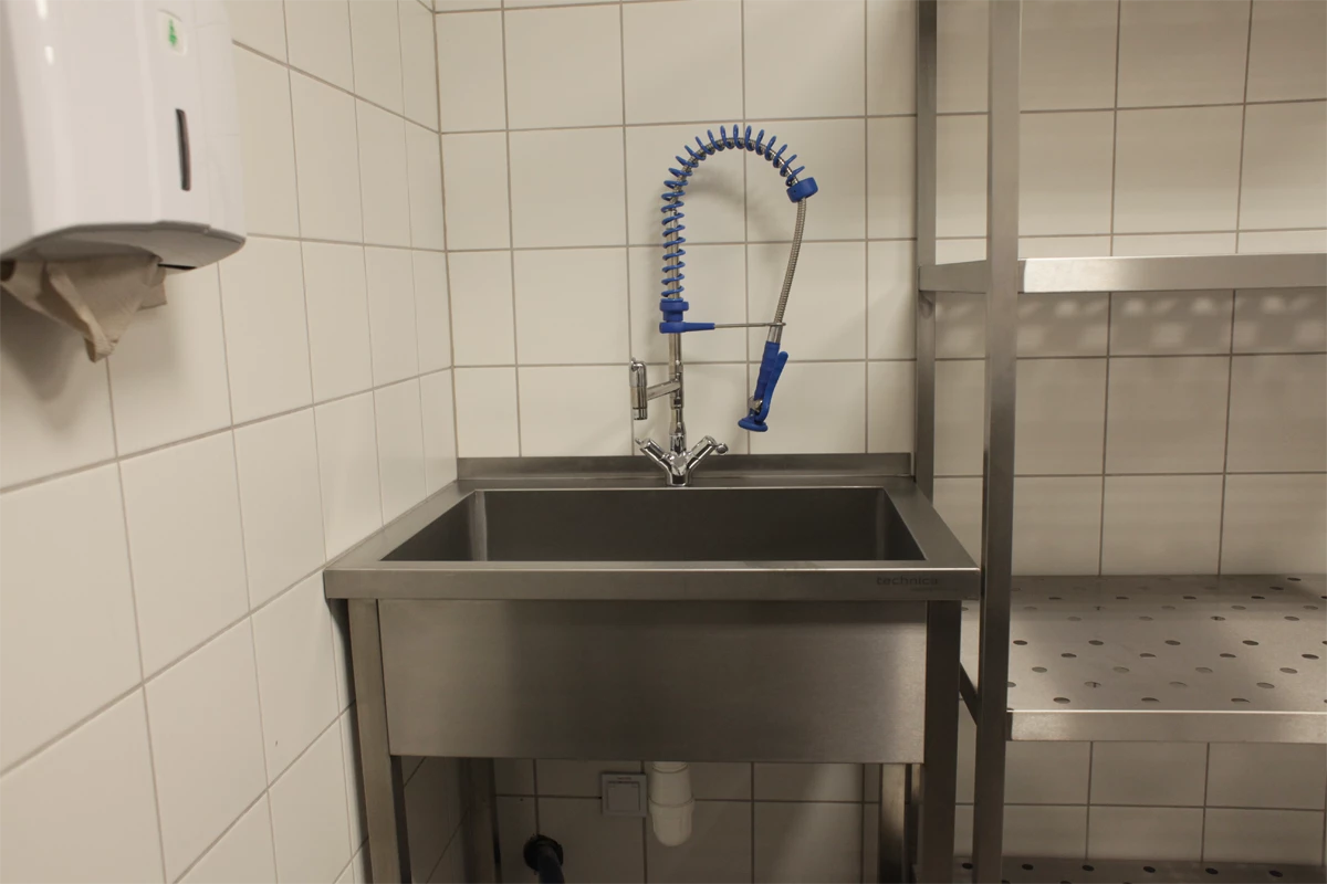 Zlew z baterią gastronomiczną, umieszczony na roboczym blacie kuchennym. Obok zlewu znajduje się dozownik papierowych ręczników, a w tle widać stalowy regał.