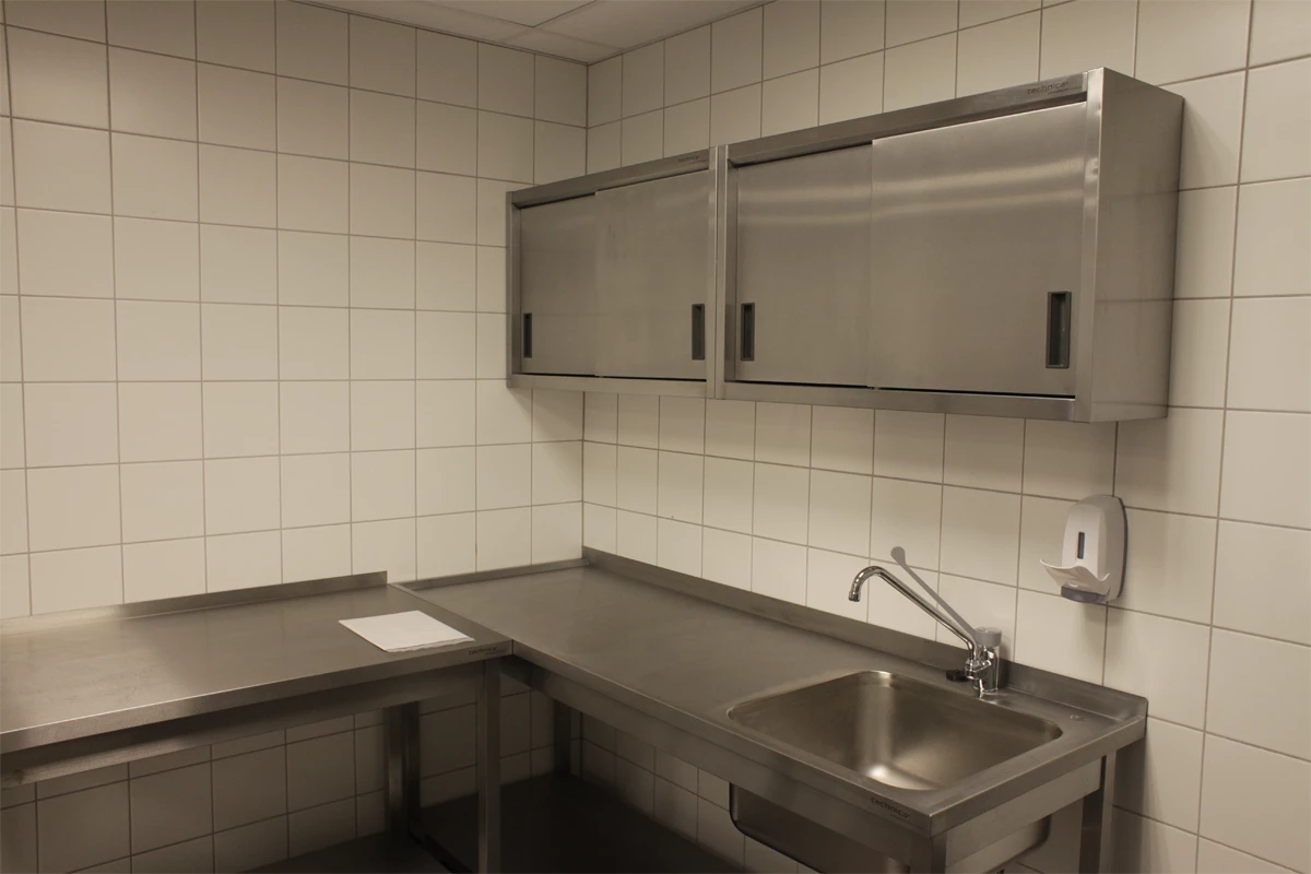 Kompleksowe stanowisko robocze w kuchni gastronomicznej wyposażone w zlew, stalowy blat i wiszące szafki Technica, na tle jasnych płytek – ergonomiczne i łatwe w utrzymaniu czystości.