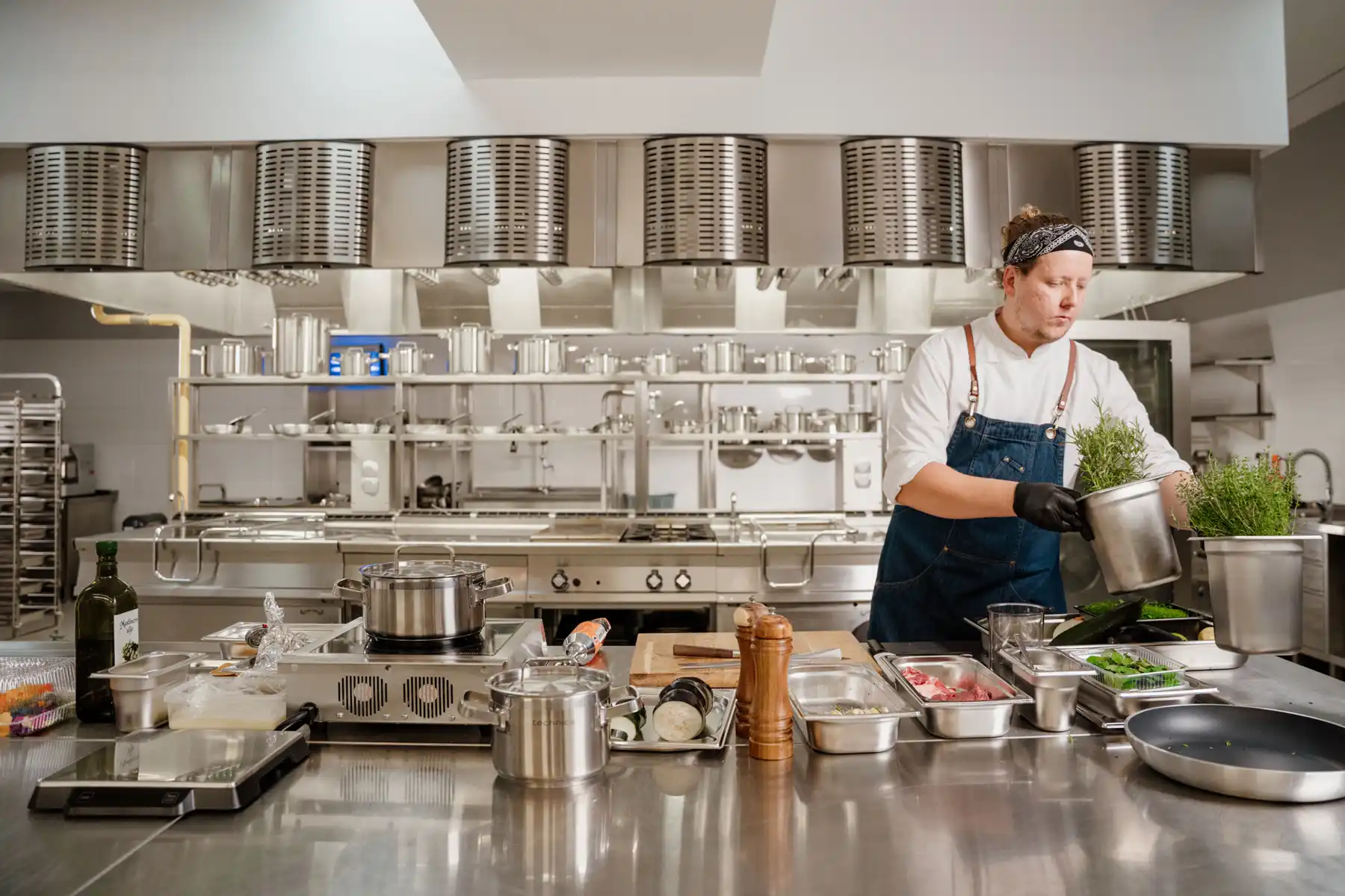 Profesjonalna kuchnia gastronomiczna ze stali nierdzewnej; kucharz Tomasz Rymisz w fartuchu roboczym przygotowuje składniki i świeże zioła na stanowisku roboczym.
