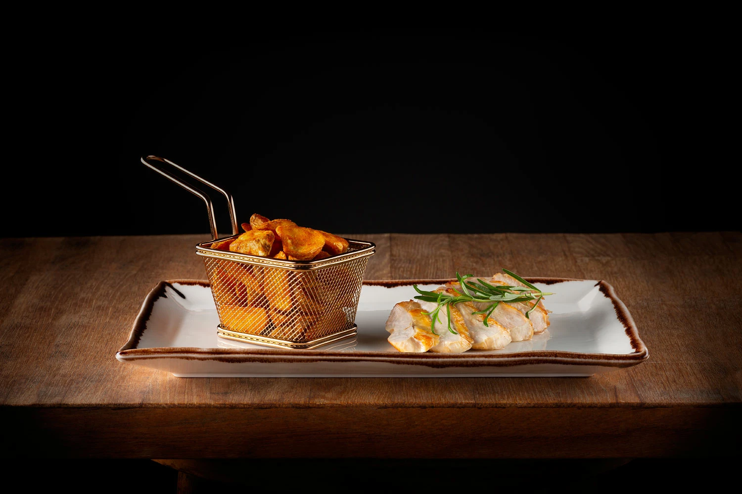 Kompozycja restauracyjna na prostokątnym talerzu – chrupiące chipsy serwowane w metalowym koszyku oraz plastry grillowanego kurczaka udekorowane świeżymi ziołami. Eleganckie i kreatywne podanie klasycznego dania w nowoczesnej odsłonie.