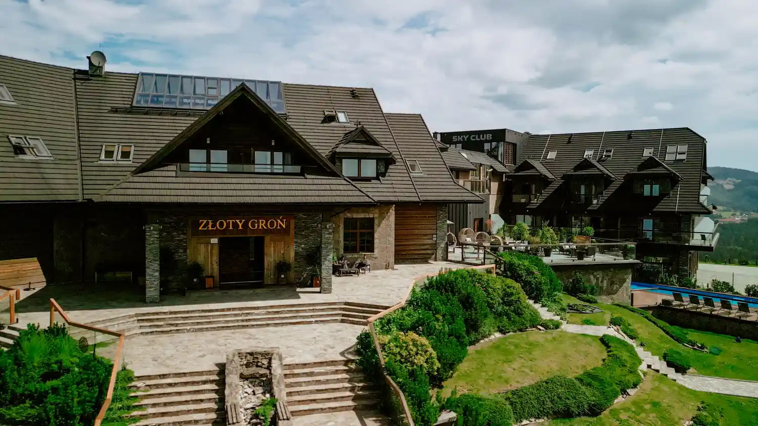 Widok z zewnętrz na Złoty Groń – kompleks hotelowo-restauracyjny w górskim otoczeniu z tarasami i zielenią.